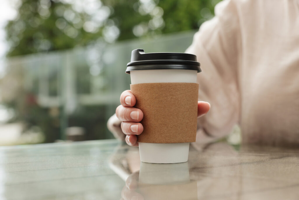 A close-up on a person's hand going to grasp a hot beverage cup with a protective paper sleeve. The person is sitting at a table with a slight reflection.