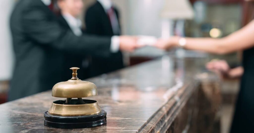 A hotel service bell sitting on a counter while a group of concierge workers greet a guest in the background.
