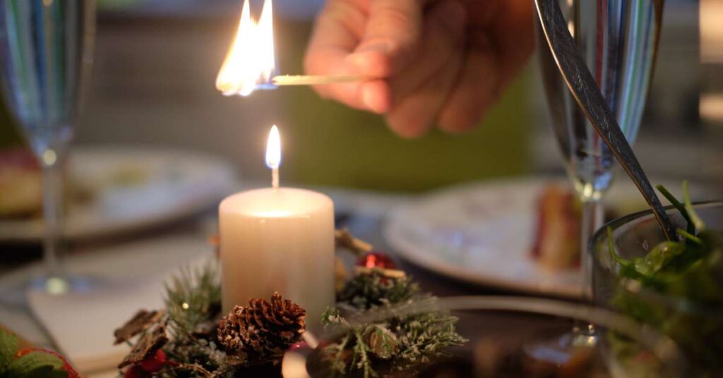 A focus on a hand lighting a candle centerpiece on a table with greenery, champagne glasses, plates, and tablesettings.