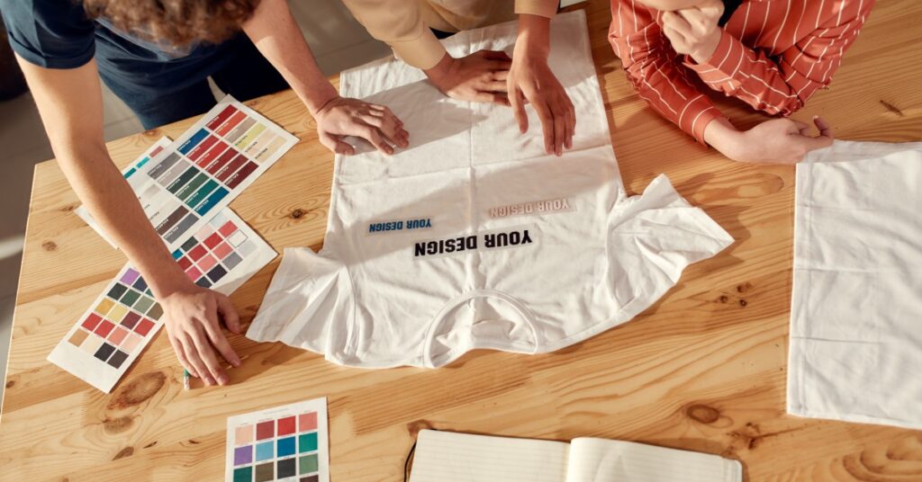 A group of three people designing a white T-shirt. The T-shirt lies flat on the table among color charts and font options.