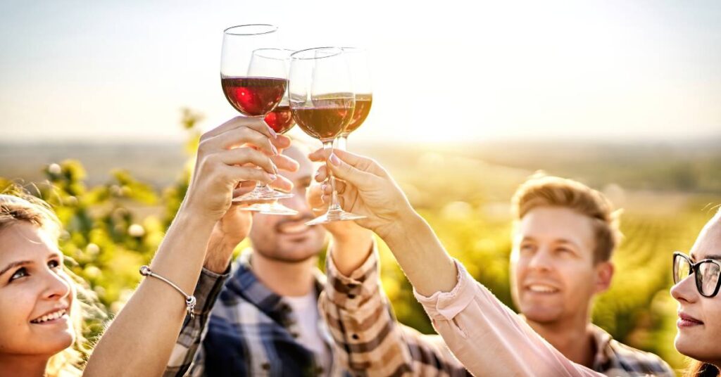 Two men and two women are drinking wine outside on a sunny day. They are holding up their glasses and toasting one another.