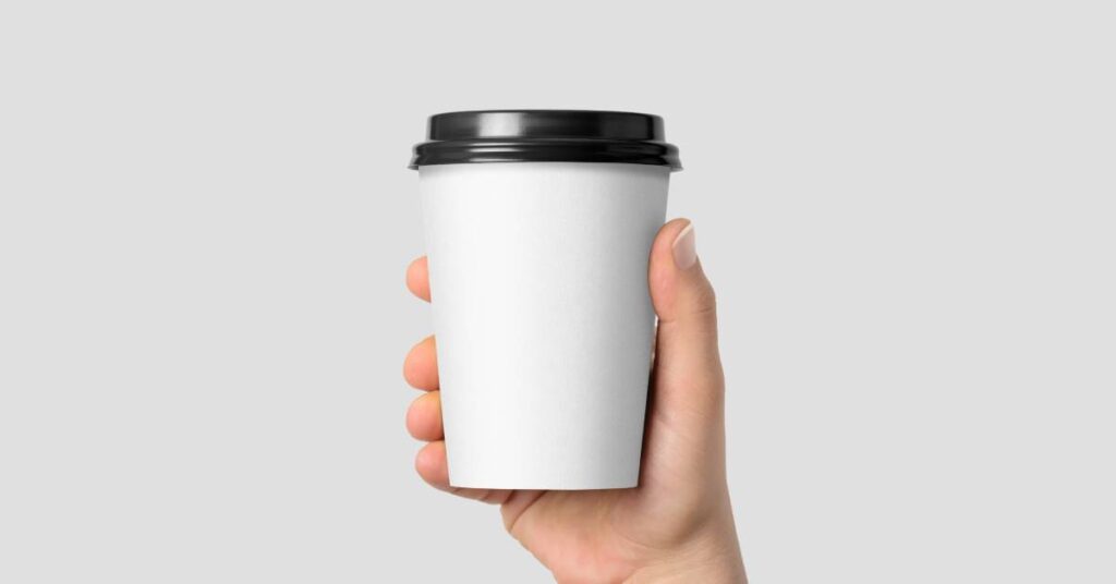 A close-up view of a hand holding a disposable coffee cup on a light gray background. The cup is white and has a black lid.