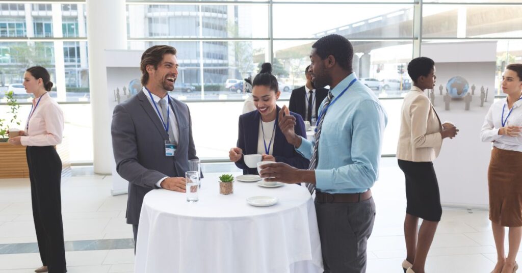 A group of individuals in professional attire socializing while standing around a cocktail table in a bright room.