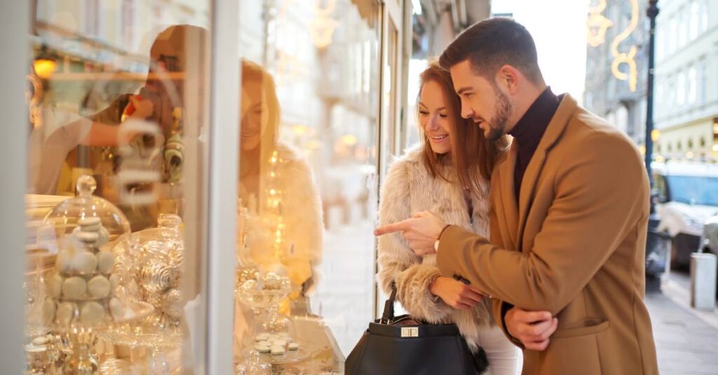 A man and a woman wearing winter coats and walking outside. They admire a store's holiday-themed window display.