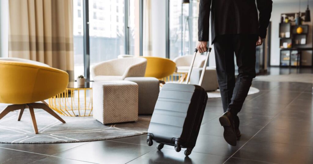 A businessman wearing a black suit is pulling a black rolling suitcase through a hotel lobby with colorful furniture.