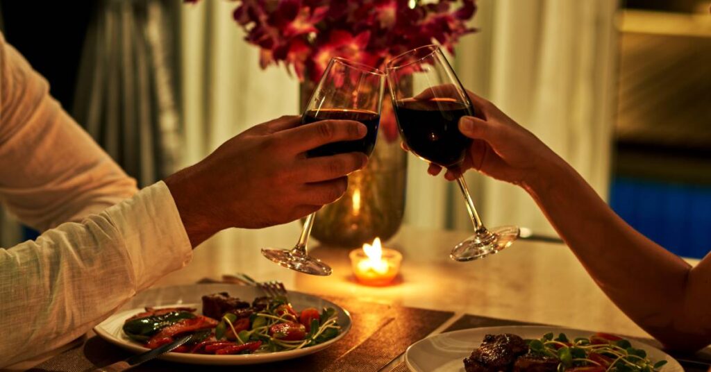 Two people eating a meal at a table with a candle and a vase of flowers. They are toasting with glasses of red wine.