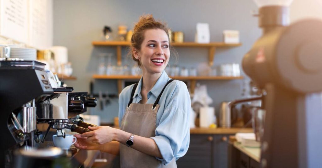 A woman in a blue shirt and a taupe apron using an espresso machine to make a beverage in a coffee shop.