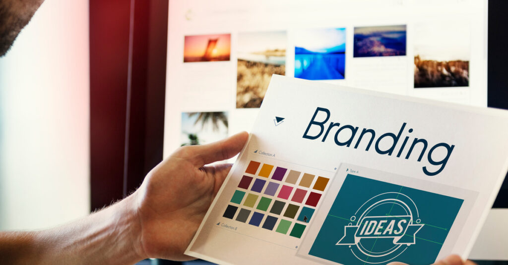 A man looking at images on a desktop computer. He is holding a piece of paper that has color swatches under the word "Branding."