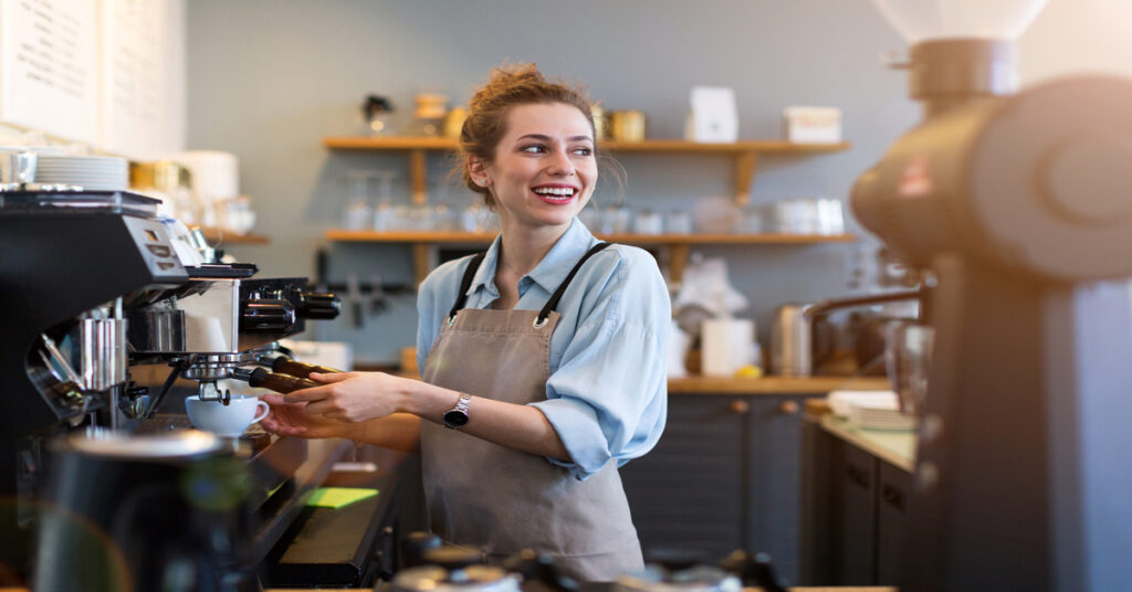 A smiling woman wearing a blue shirt and brown apron using a coffee machine to make a beverage in a coffee shop.