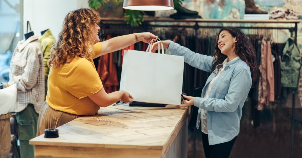 A female employee of a clothing store is standing behind a counter and handing a white shopping bag to a customer.