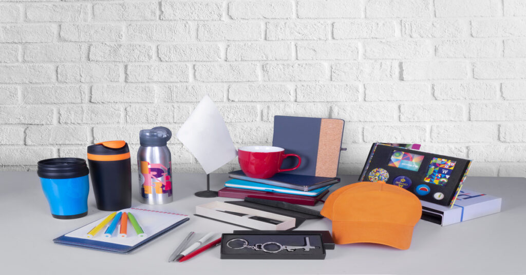 An assortment of promotional products, including a mug, hat, notebooks, pens, and water bottles, on a white brick background.