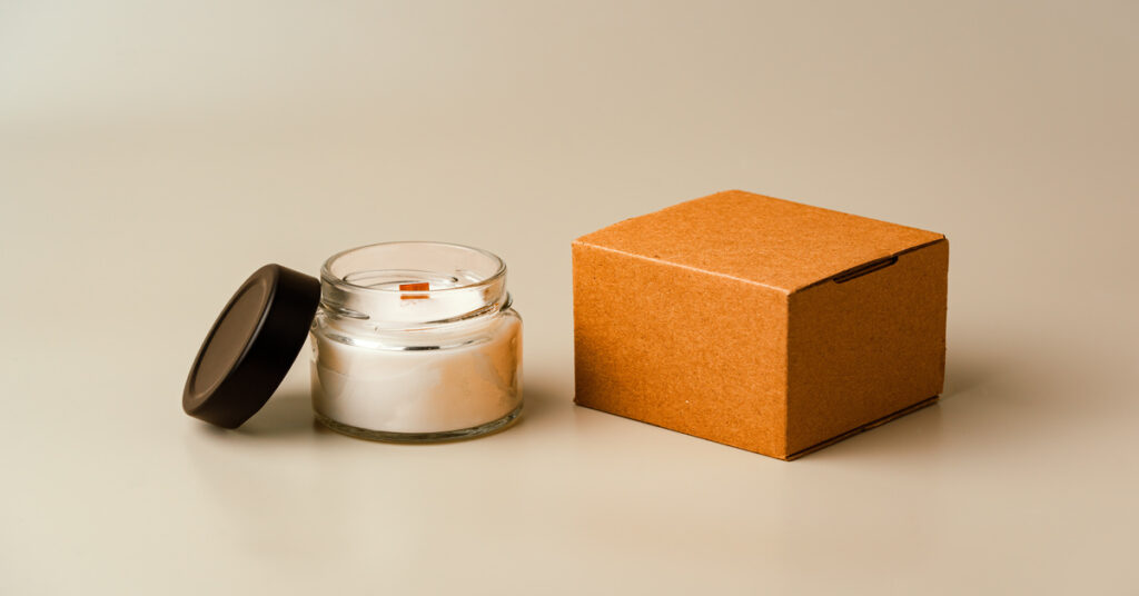 A glass candle jar and a cardboard box set against a neutral background. The black lid of the candle is resting against the jar.