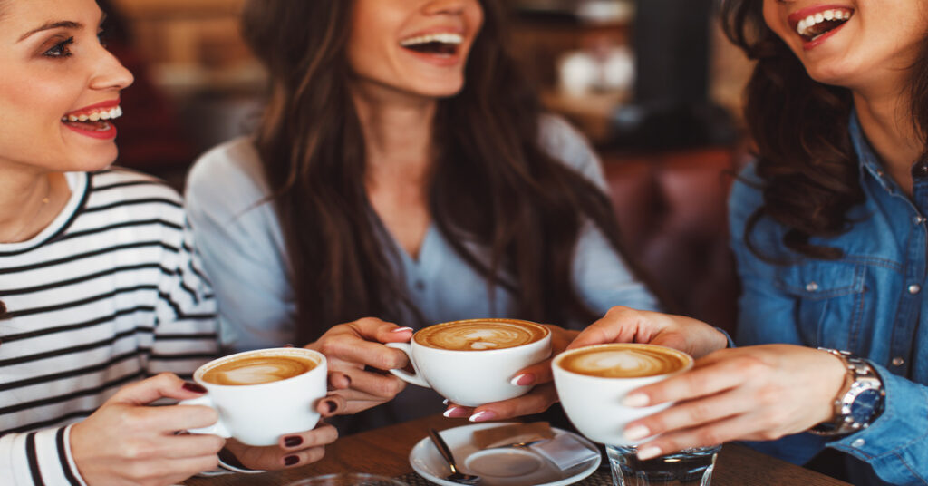 A group of three smiling women sitting next to each other at a table and drinking coffee in white mugs.