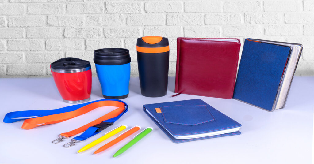 An assortment of promotional products, including notebooks, drinkware, pens, and lanyards, against a white brick wall.