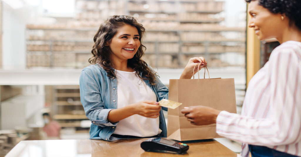 A smiling female customer holding a brown paper bag while handing her credit card to a store employee.
