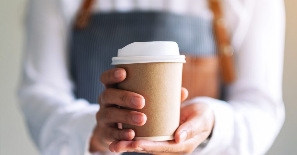 An individual wearing a blue apron and a white shirt holding a brown paper coffee cup with a white lid.