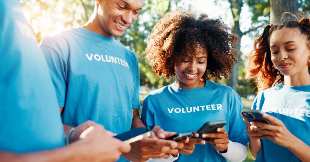 A group of four young adults in blue T-shirts that say "Volunteer" looking at their cell phones while standing outside.