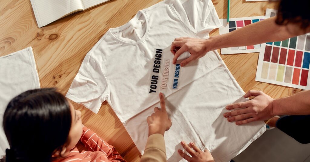 An aerial view shows a group of people using colorful samples labeled "Your Design" to create a custom white T-shirt.