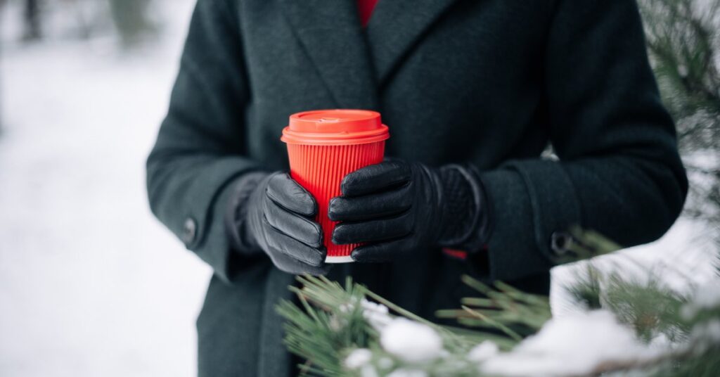 A person in a black coat and leather gloves holds a red disposable coffee cup outside on a snowy day.