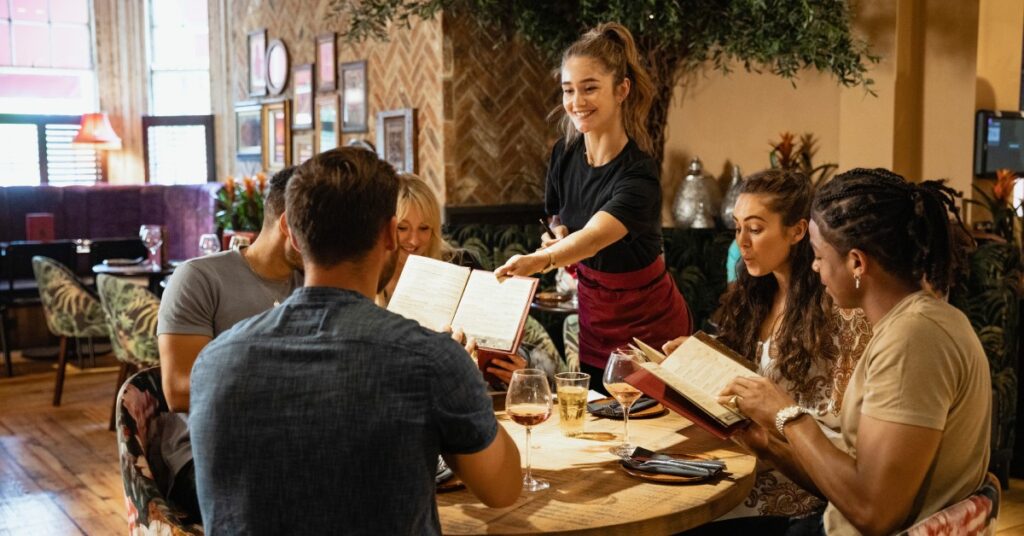 Five young people sit at a circular table at a restaurant while a smiling female server hands them menus.