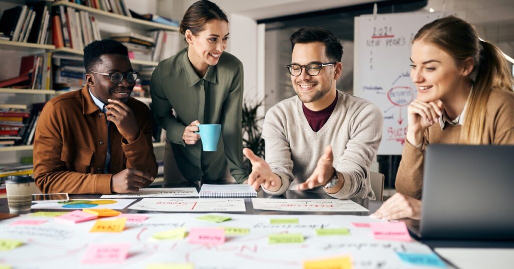 Two male and two female colleagues hold a meeting, reviewing a large sheet of paper covered in sticky notes.