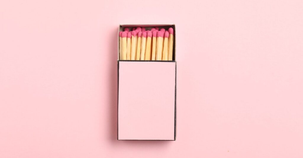 An overhead view of an open matchbox with pink-tipped matches neatly arranged, set against a soft pink background.