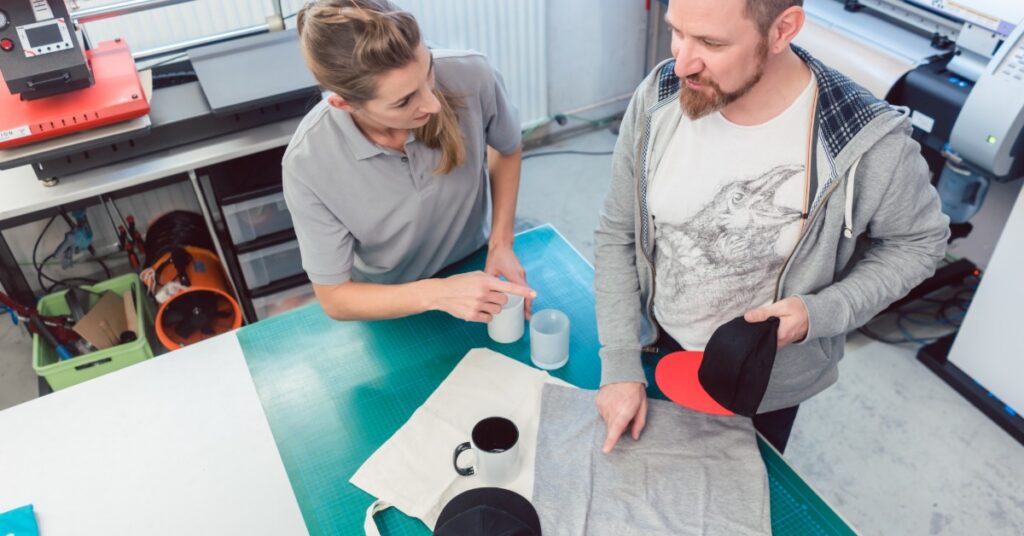 Two people reviewing custom apparel and mugs on a worktable with hats and printing equipment in a workshop setting.