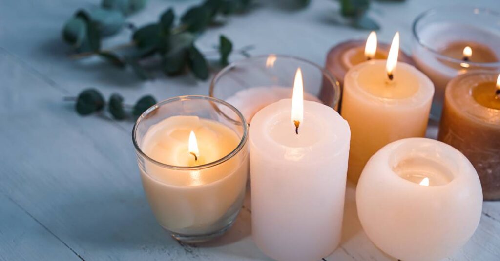 A group of lit pillar and votive candles on a white wooden table with soft greenery in the background. File Name: MarylandMatch-429998-Candles-Wooden-