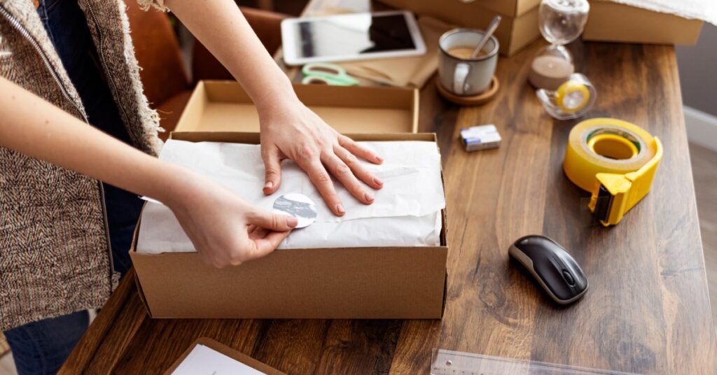 A person sealing a tissue-wrapped package in a cardboard box on a wooden desk with packing supplies nearby.