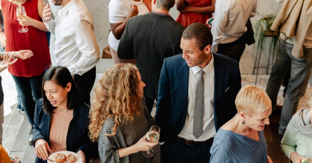Professionals mingle at a crowded corporate networking event, holding drinks and chatting indoors together.