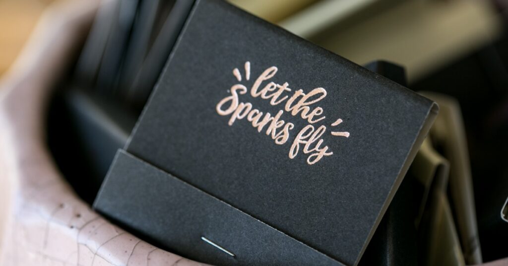 Black wedding matchbooks in a decorative bowl with rose gold “let the sparks fly” lettering and blurred matches behind.