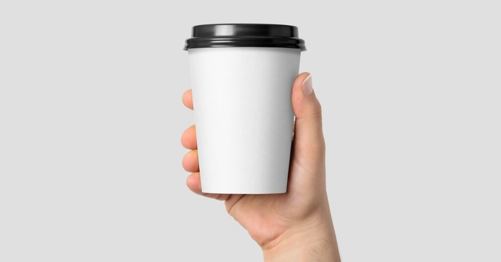 A hand holding a plain white disposable coffee cup with a black lid against a light gray background.