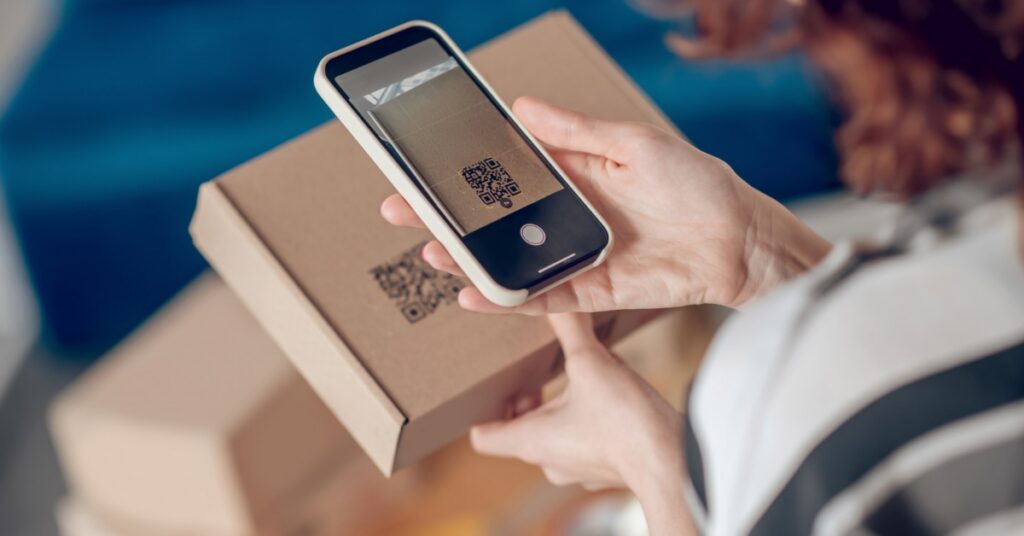 Person scanning a QR code on a cardboard shipping box with a smartphone, demonstrating contactless product access.