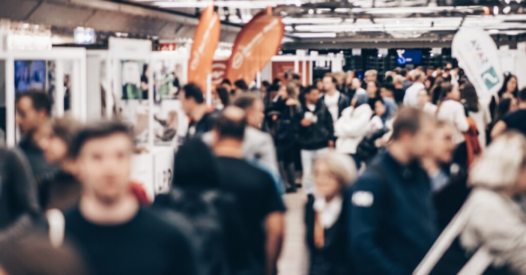 Crowded trade show floor filled with attendees walking among exhibitor booths, displays, and colorful promotional banners.