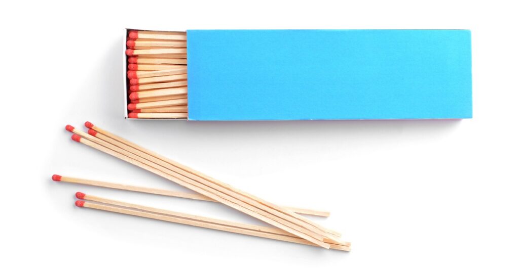 Long wooden matches with red tips lie below an open bright blue matchbox filled with additional matches.