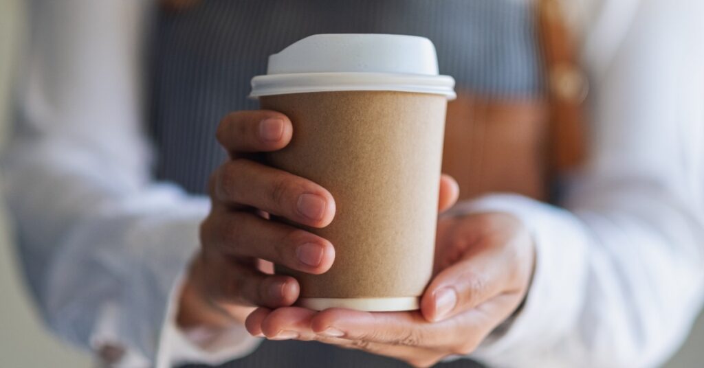 Person in an apron holds a plain brown paper coffee cup with a white lid in both hands against a blurred background.