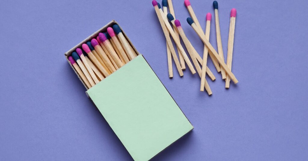 Open green matchbox filled with wooden matches with colorful tips, with loose matches scattered on a purple background.