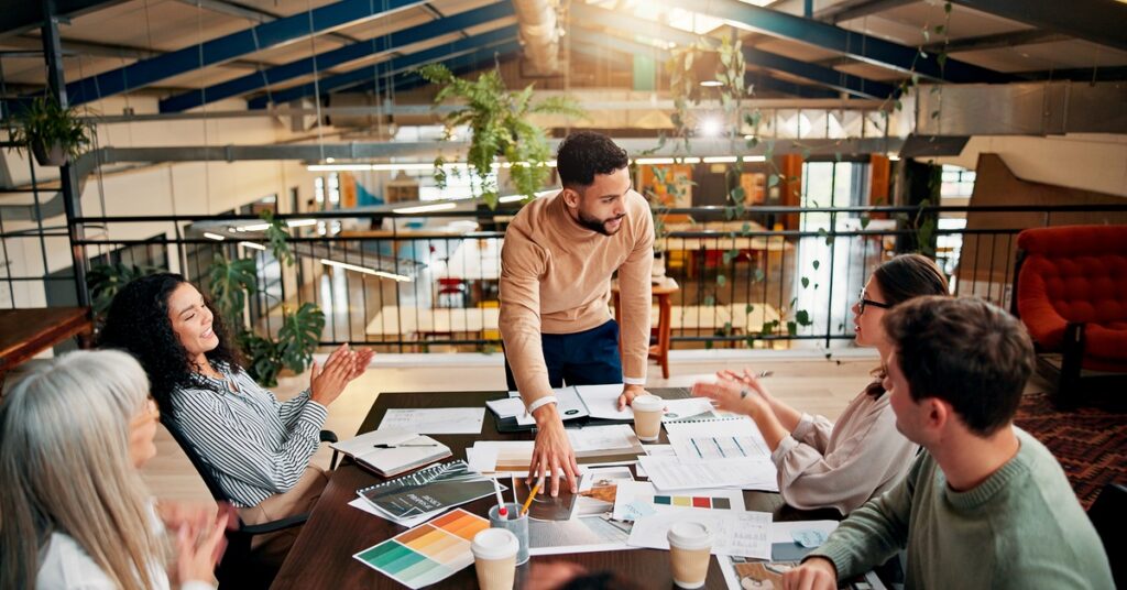 Five coworkers discuss branding ideas around a conference table covered with charts and color samples in a modern office.